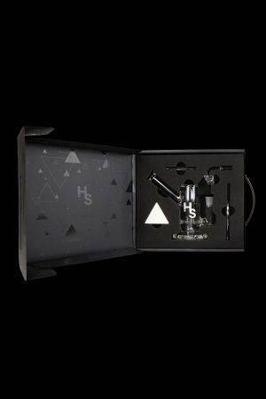 Open black gift box displaying cannabis smoking accessories, including a glass pipe, vaporizer, and tools, with a geometric pattern and "Hs" logo.