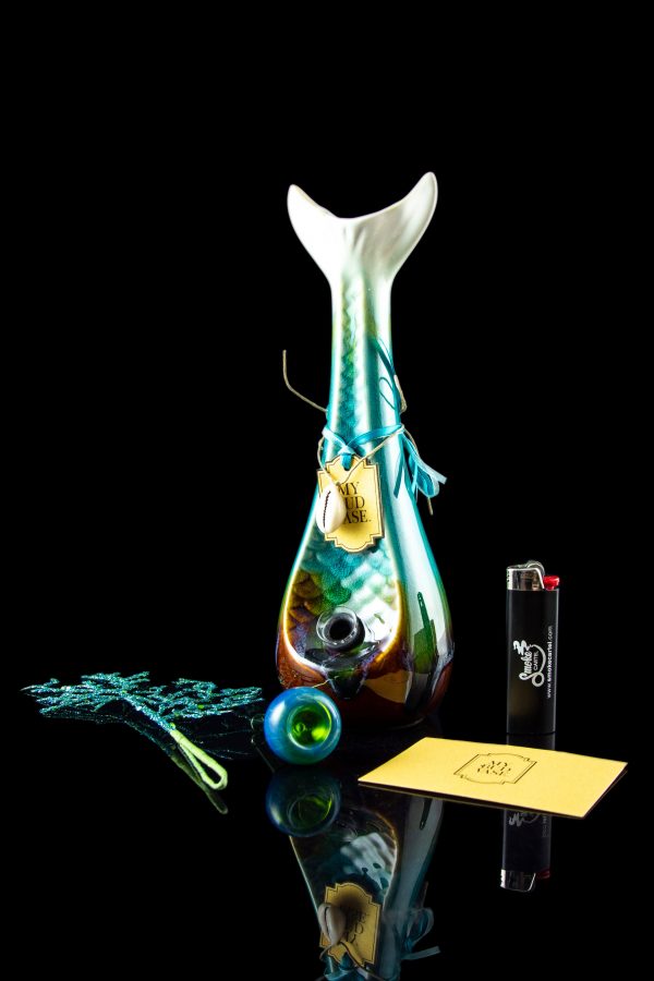 A vibrant, iridescent glass water pipe (bong) with blue and teal swirls is displayed alongside cannabis accessories: a small glass marble, a rolling paper, a lighter, and a small container with plant matter.