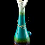 A vibrant green and blue glass water pipe with a rounded base and intricately sculpted neck, tied with natural twine, against a black background.