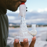 A person holds and inhales from a rocket-shaped glass water pipe, with a blurred landscape background.