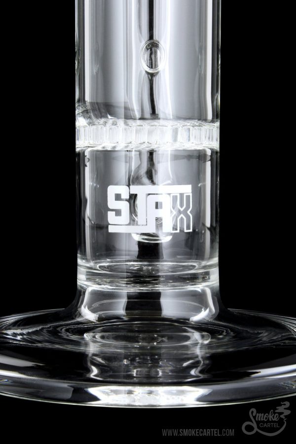 Close-up of a clear glass water pipe with “STAX” etched on the side, filled with water and reflecting on a dark surface.