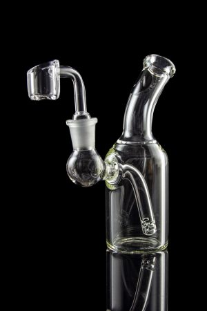 Clear glass dab rig with quartz banger, reflecting on a dark surface.