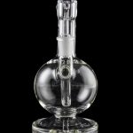 Clear glass water pipe (rig) with spherical body and intricate internal pathways, reflecting on a dark surface.