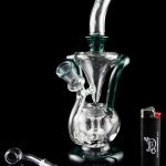A clear glass water pipe (rig) with a unique sculptural design, alongside cannabis oil concentrate dab tools and a lighter, all reflected on a black surface.