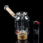 Glass water pipe with a wooden mouthpiece and quartz insert, displayed with a lighter on a reflective surface.