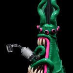 Green glass water pipe shaped like a monster with a pink tongue and exposed teeth, accompanied by a quartz banger.