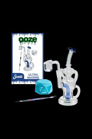 A studio shot of a clear glass water pipe ("rig") alongside cannabis accessories including a dab tool, silicone container, and product packaging for "Ooze Swell Ultra Marine."