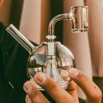 A person holds a clear glass bubbler/water pipe, often used for cannabis consumption."