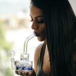 A woman with long dark hair inhales from a small, clear glass rig with blue accents, likely for cannabis consumption.