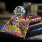 A glass mushroom pipe rests on a patterned bandana draped over stacked vintage books, illuminated in a dimly lit, moody setting.