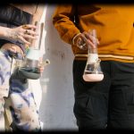 Two people holding glass bongs, partially visible against a textured wall.